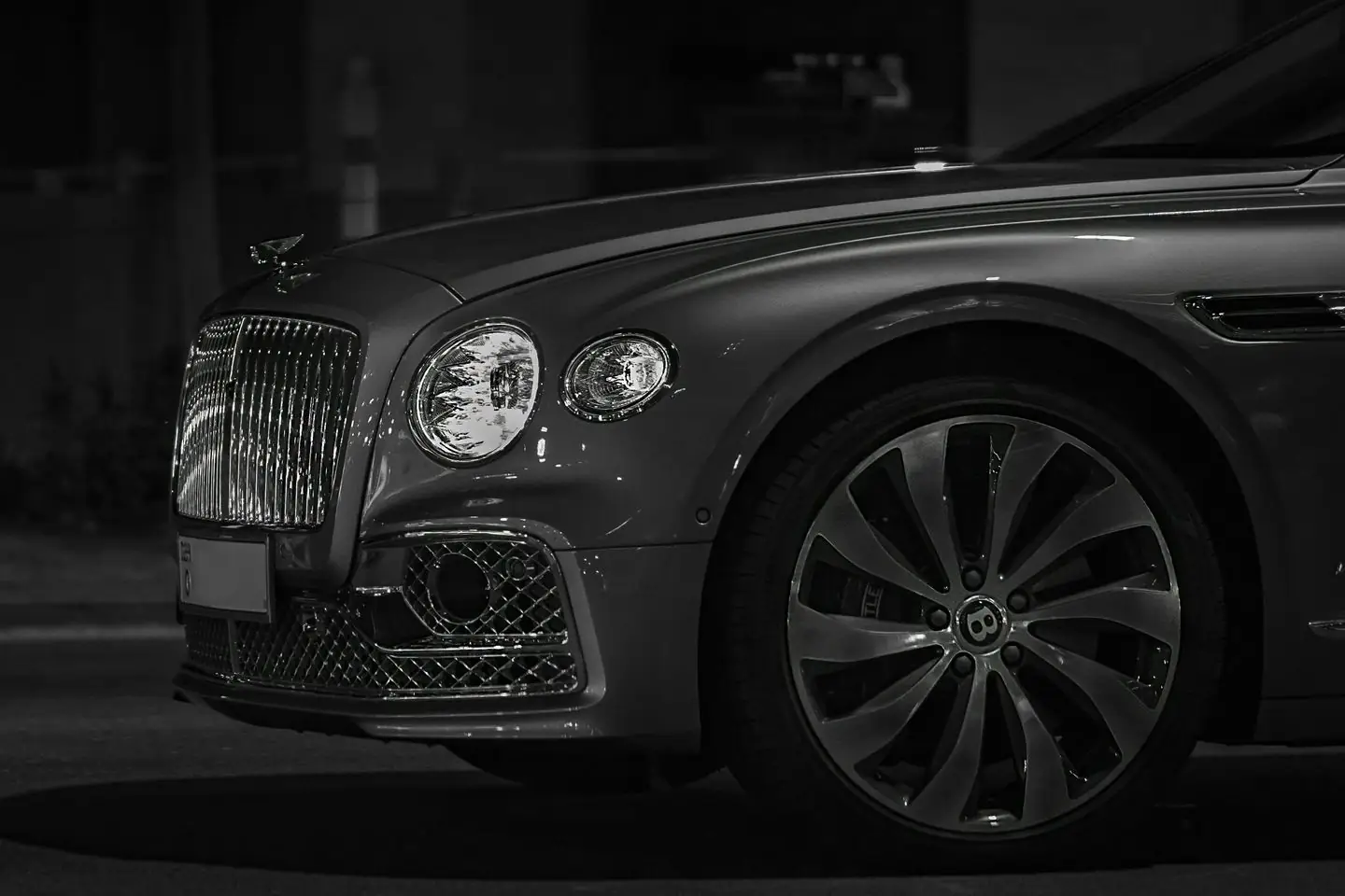 The front of a dark grey Bentley sedan at night, its grille and headlights in a low side light
