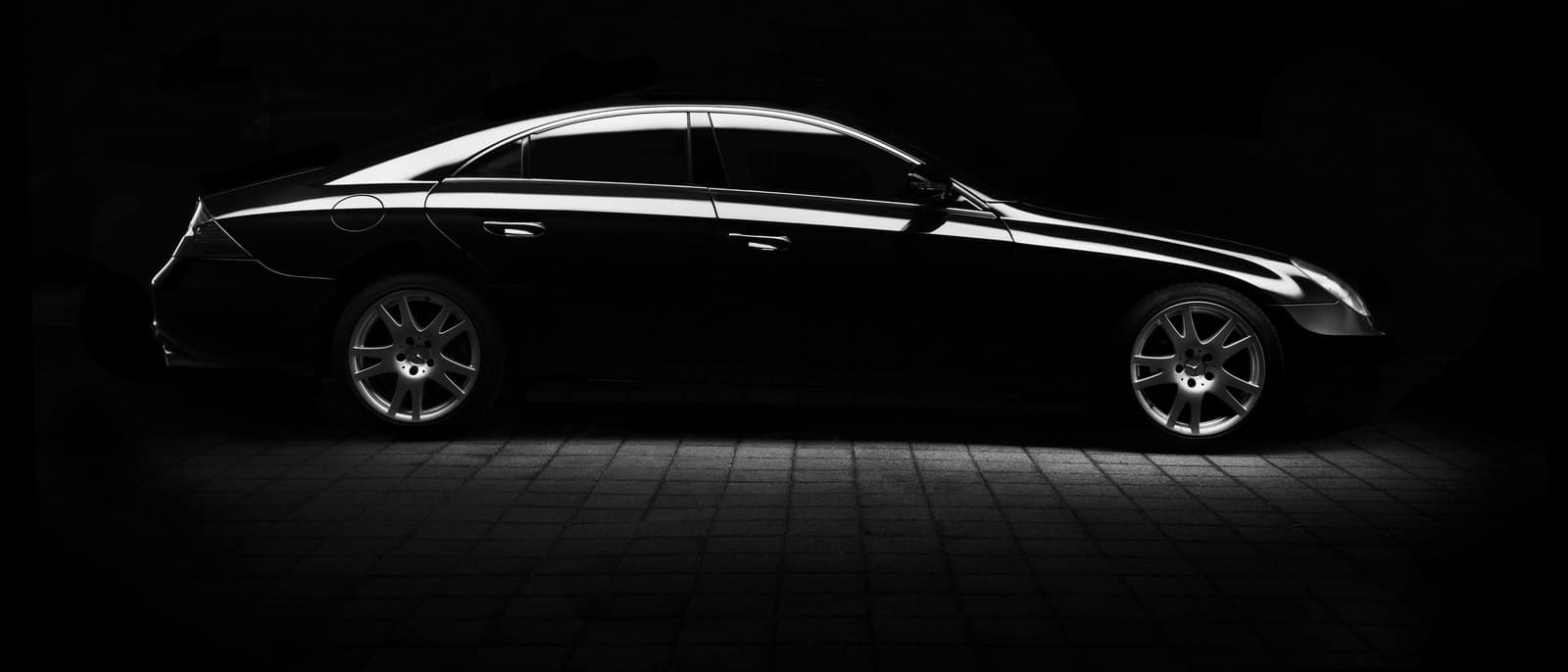 A black luxury sedan in profile against a black background, its roofline and wheels picked out by a low side light