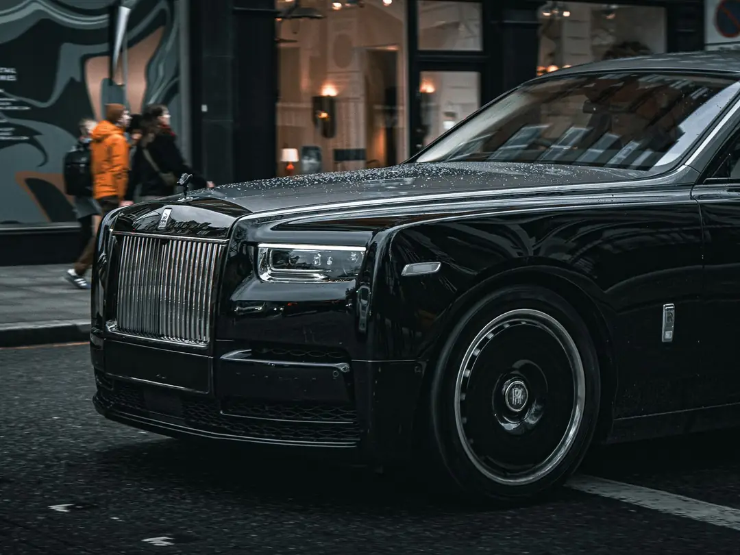 The front of a black Rolls-Royce Phantom on a wet city street, people walking past the shopfronts behind it
