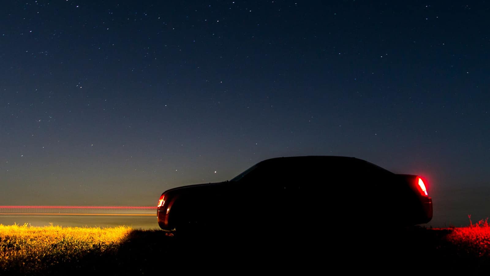 A dark sedan in silhouette under a starry evening sky, its taillights lit red